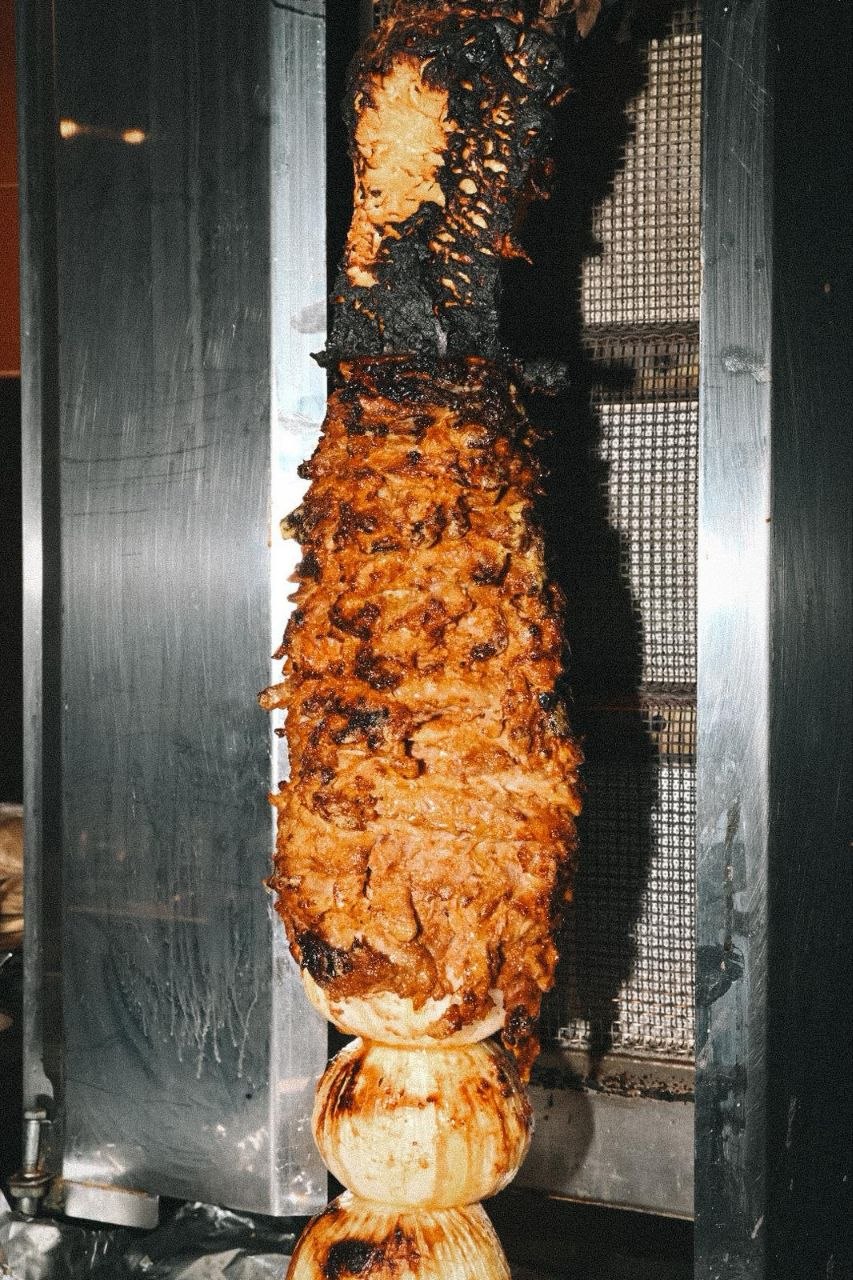 Trompo de carne adobada coronado por una piña, girando sobre el fuego en la cocina de ETL. La piña gotea su jugo sobre las capas de carne al pastor.