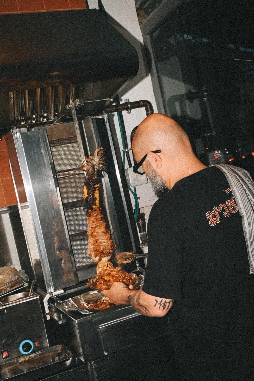 Juan Pescador frente a un trompo de pastor en cocina, abrigo blanco, gesto concentrado.