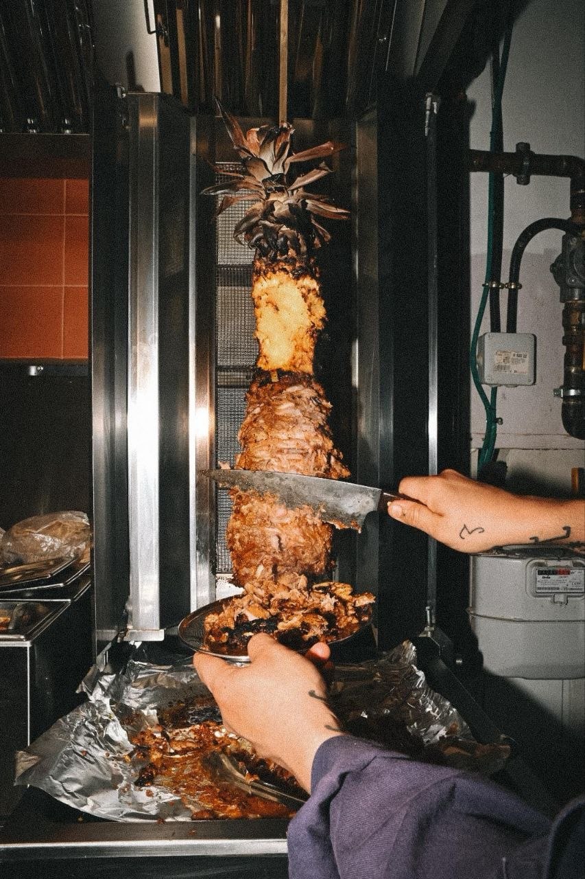 Mano del taquero cortando láminas finas de carne adobada del trompo encendido; el cuchillo largo entra horizontal, las virutas caen sobre una tortilla pequeña.
