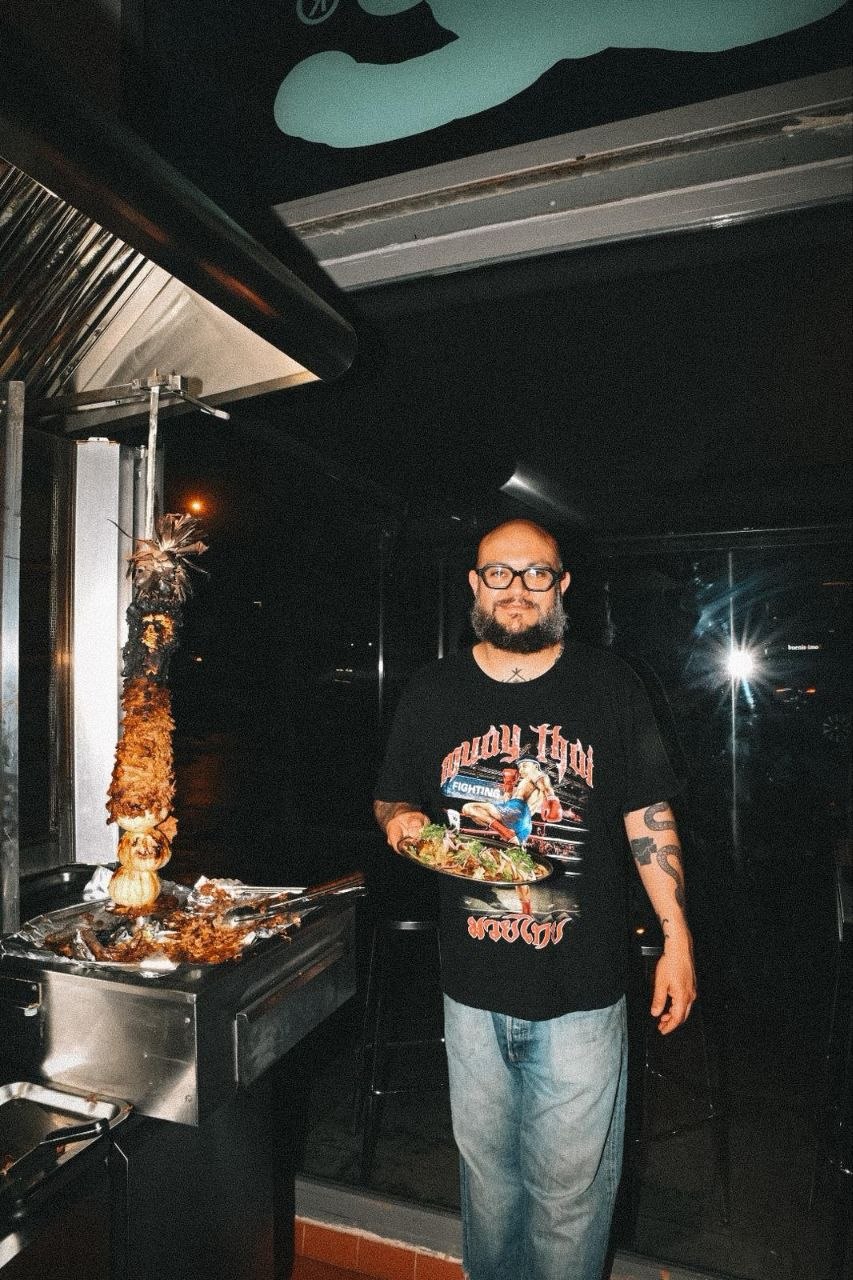 Juan Pescador en cocina, junto a un plato de tacos. Camiseta gris, fondo neutro.