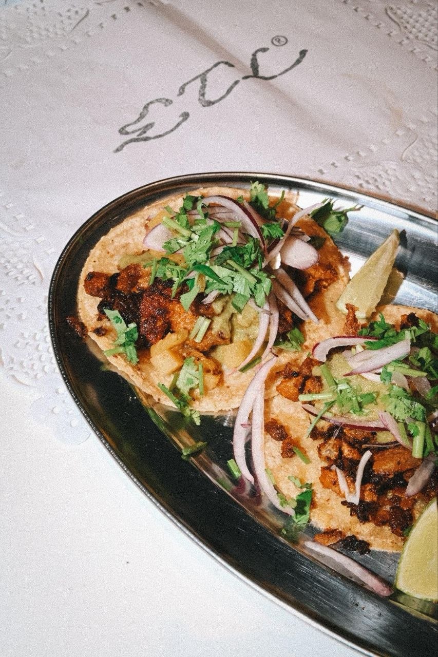 Pequeña tabla con tres tacos servidos sobre una servilleta de papel calado, con la firma cursiva ETL escrita a mano debajo.